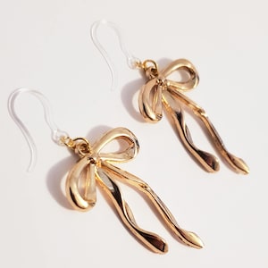 May include: A pair of gold-colored bow earrings with clear plastic ear wires. The bows have a textured, metallic finish and flowing ribbon-like tails. The earrings are displayed against a white background.
