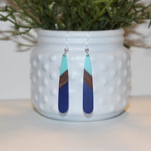 May include: A pair of teardrop-shaped earrings with a blue, brown, and turquoise color scheme. The earrings are made of resin and have a silver hook.