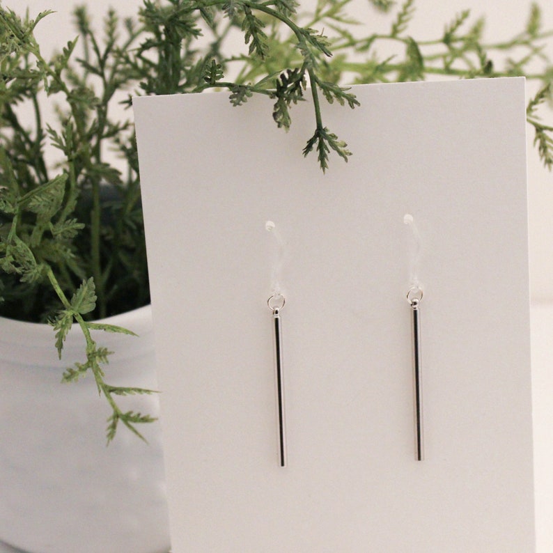 May include: A pair of silver dangle earrings with thin, straight bars hanging from small loops. The earrings are displayed on a white card.