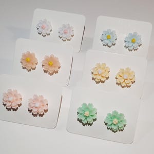 May include: A collection of floral stud earrings in various colors, including white, pink, yellow, blue, and green. Each pair of earrings is displayed on a white card. The earrings have a detailed, layered petal design with a contrasting center.