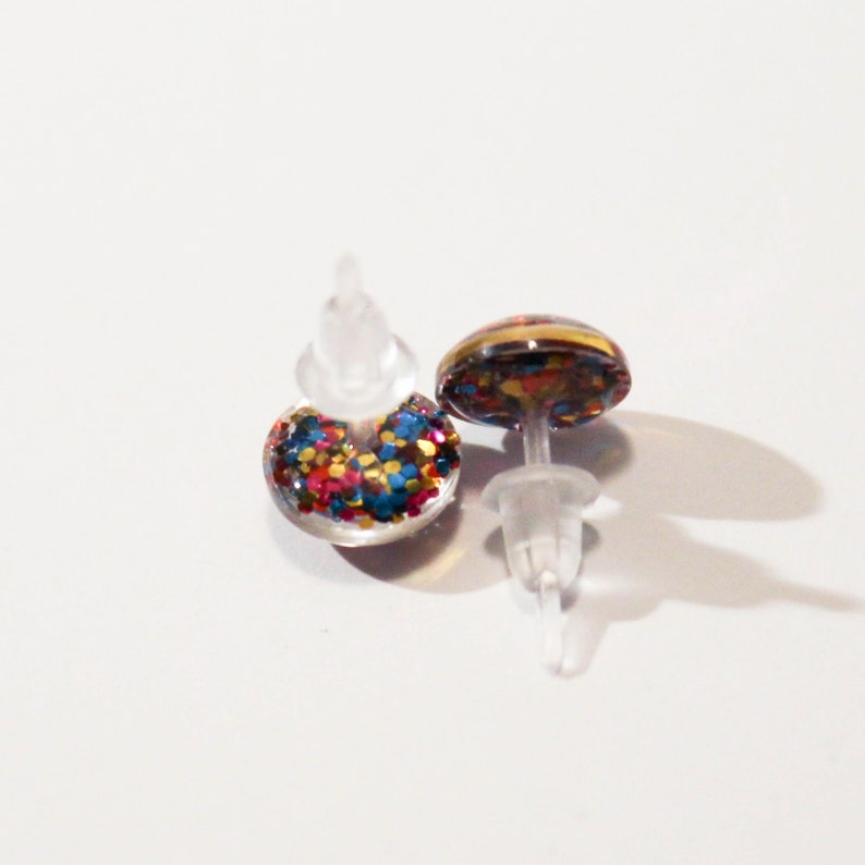 May include: A pair of stud earrings with a clear plastic post and a round, colorful glitter design. The glitter is red, blue, gold, and pink.