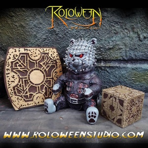 Puede incluir: Un oso de peluche gris y negro con una cabeza plateada puntiaguda y ojos rojos. El oso está sentado sobre una superficie marrón con dos cajas de rompecabezas doradas. El texto "ROLOWEEN STUDIO" es visible en el fondo.