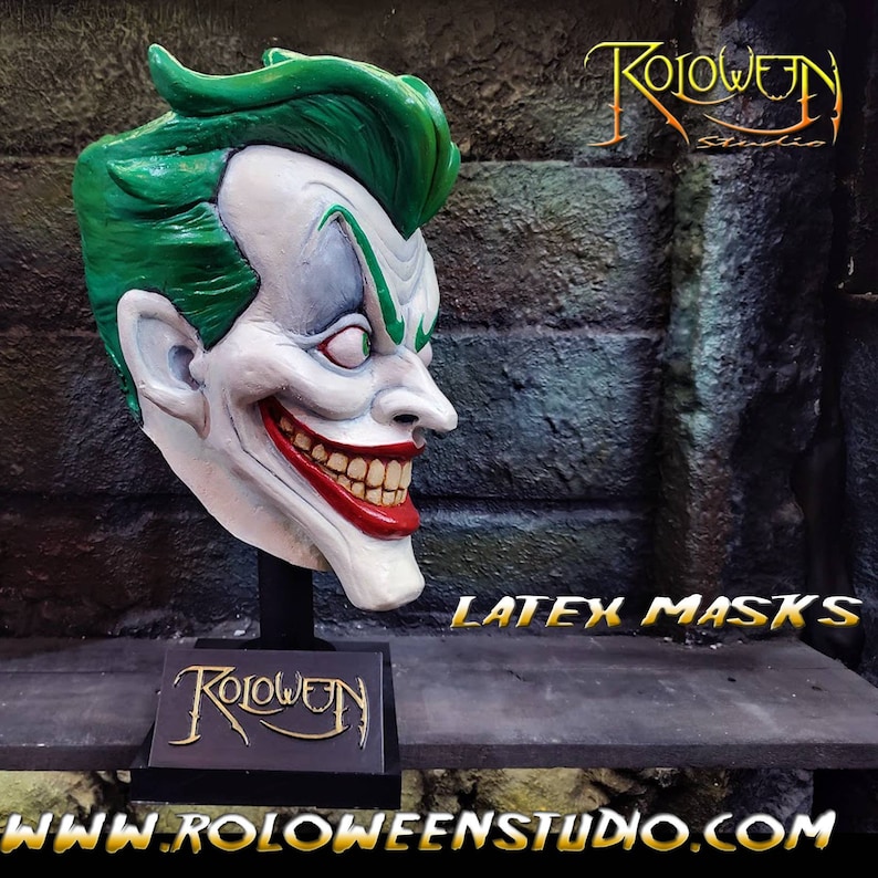 May include: A green and white latex Joker mask with a wide, toothy grin. The mask is on a black stand with the words "Roloween Studio" in gold lettering. The text "LATEX MASKS" is visible in gold lettering on a dark surface. The website address "www.roloweenstudio.com" is visible in gold lettering at the bottom of the image.