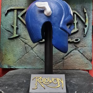 May include: Blue and white plastic helmet with a wing design on the side. The helmet is on a black stand with the word "Roloween" written on the base.