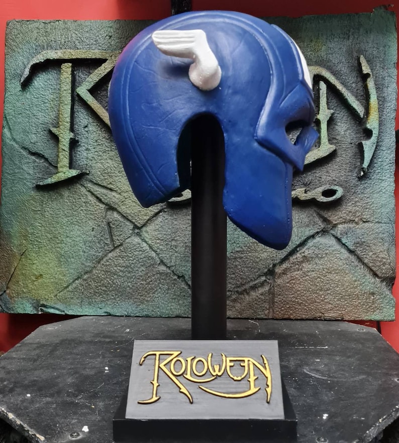 May include: Blue and white plastic helmet with a wing design on the side. The helmet is on a black stand with the word "Roloween" written on the base.