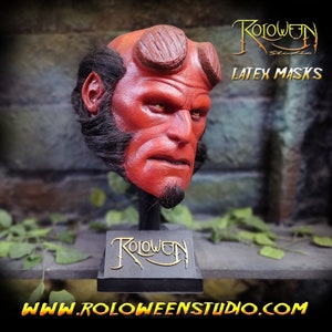 May include: A red latex mask of the character Hellboy, with horns and a beard, on a black display stand. The stand has the text "Roloween Studio" in gold lettering. The image also includes the text "LATEX MASKS" and "WWW.ROLOWEENSTUDIO.COM".