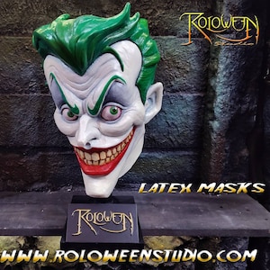 May include: A green and white latex mask of the Joker, with a wide, toothy grin. The mask is on a black stand with the words "Roloween Studio" in gold lettering. The text "Latex Masks" is visible in the background.