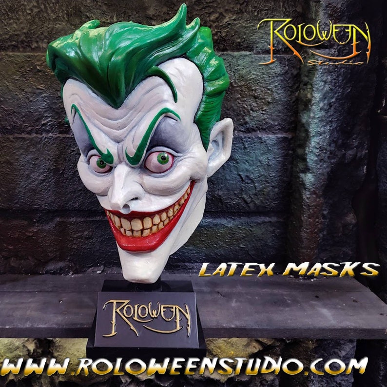 May include: A green and white latex mask of the Joker, with a wide, toothy grin. The mask is on a black stand with the words "Roloween Studio" in gold lettering. The text "Latex Masks" is visible in the background.