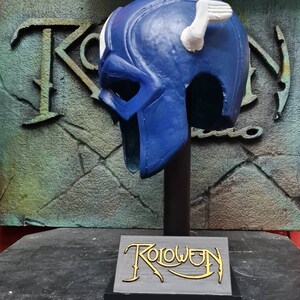May include: A blue and white plastic helmet with a white wing on top. The helmet is on a black stand with a gray base that says "Roloween" in gold lettering.