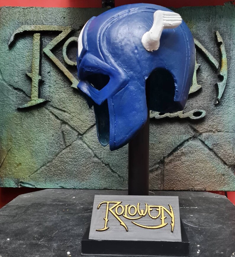 May include: A blue and white plastic helmet with a white wing on top. The helmet is on a black stand with a gray base that says "Roloween" in gold lettering.