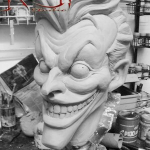 May include: A close-up of a detailed, white clay sculpture of the Joker's head. The sculpture has a wide, exaggerated smile with visible teeth and large, expressive eyes. The Joker's hair is styled in a spiky, upward fashion. The sculpture is sitting on a table with a can of paint in the background.