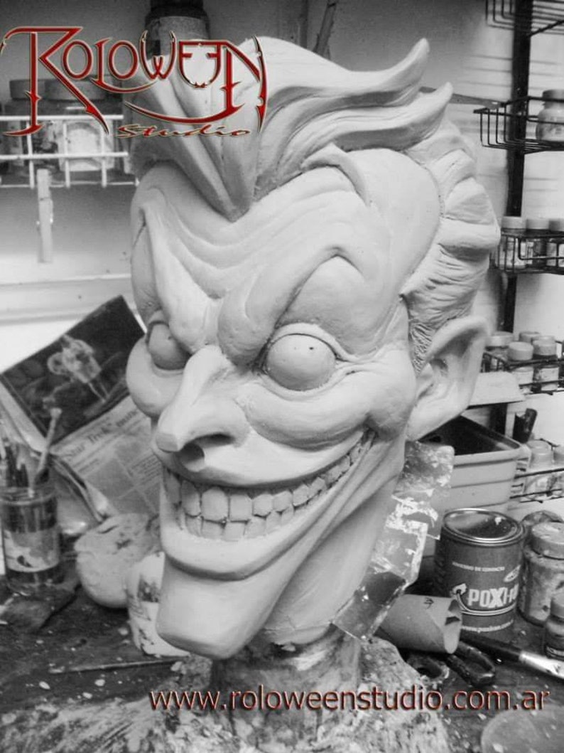 May include: A close-up of a detailed, white clay sculpture of the Joker's head. The sculpture has a wide, exaggerated smile with visible teeth and large, expressive eyes. The Joker's hair is styled in a spiky, upward fashion. The sculpture is sitting on a table with a can of paint in the background.