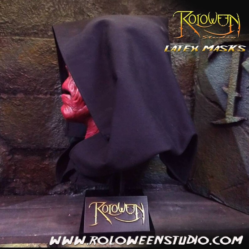 May include: A red latex mask with a black hooded robe. The mask is on a black stand with a black base. The stand has a small sign with the text "Roloween" in gold lettering. The text "LATEX MASKS" is visible in the upper right corner of the image. The text "WWW.ROLOWEENSTUDIO.COM" is visible in the lower right corner of the image.