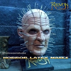 May include: A detailed latex horror mask of a character with a grid of metal pins protruding from the head. The mask is on a black stand with the text "Roloween Studio" in gold lettering.