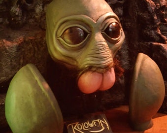 May include: A green and brown alien-like creature head with large eyes and a long tongue. The head is on a stand with the text "Roloween" on it. The head is part of a costume.