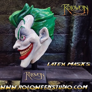 May include: A white latex Joker mask with green hair, red lips, and a wide, toothy grin. The mask is mounted on a black stand with the words "Roloween Studio" printed on it. The image also includes the text "LATEX MASKS" and the website address "www.roloweenstudio.com".