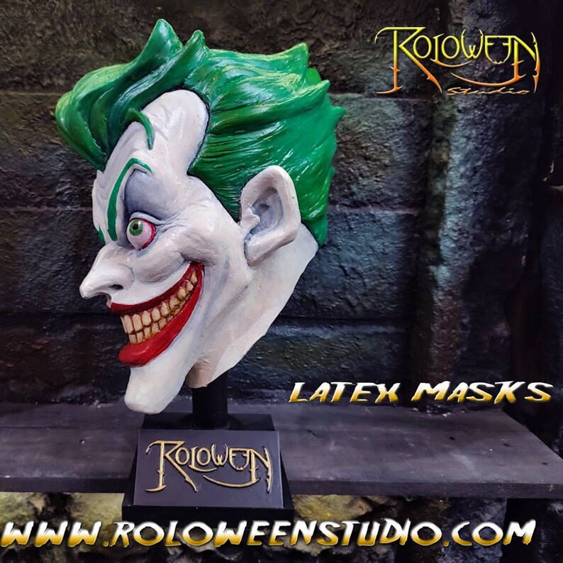 May include: A white latex Joker mask with green hair, red lips, and a wide, toothy grin. The mask is mounted on a black stand with the words "Roloween Studio" printed on it. The image also includes the text "LATEX MASKS" and the website address "www.roloweenstudio.com".
