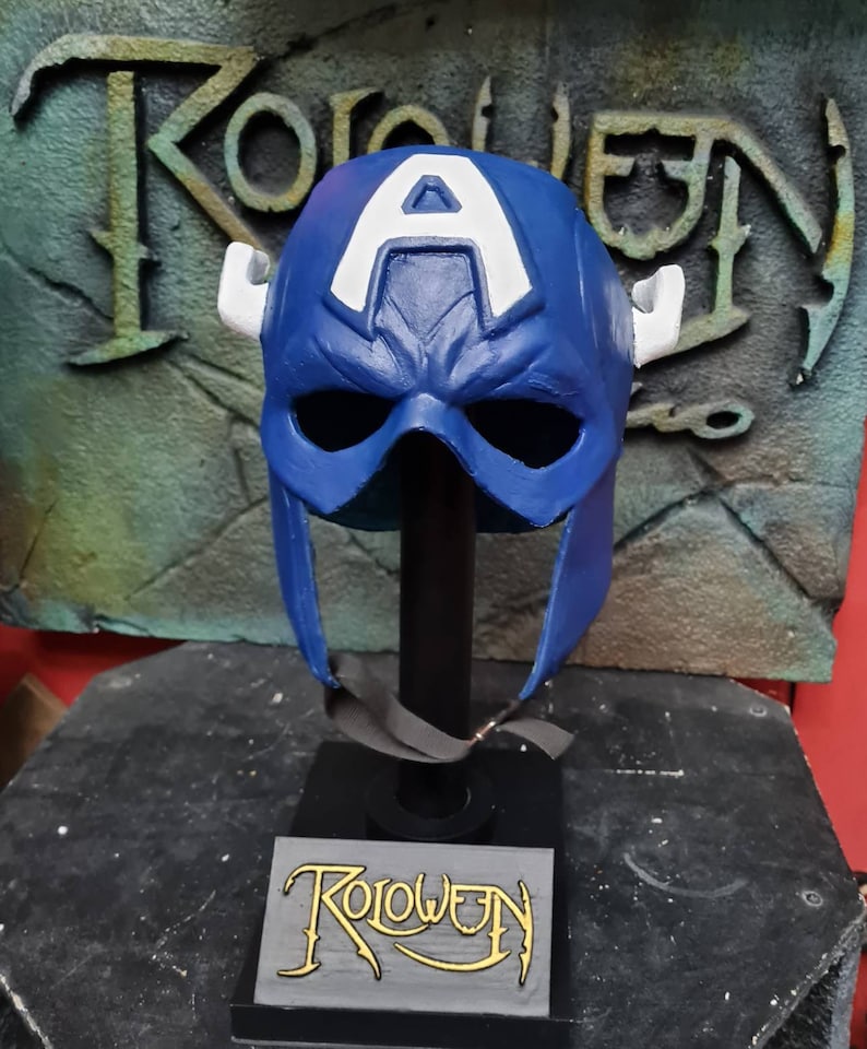 May include: A blue and white Captain America helmet with horns on the sides, displayed on a black stand with the word "Roloween" written on it.
