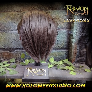 May include: A brown latex mask with short brown hair on a black display stand. The stand has the text "Roloween Studio" in gold lettering. The text "LATEX MASKS" is visible in the background.