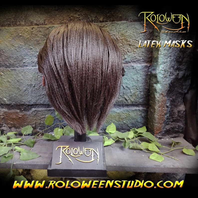 May include: A brown latex mask with short brown hair on a black display stand. The stand has the text "Roloween Studio" in gold lettering. The text "LATEX MASKS" is visible in the background.