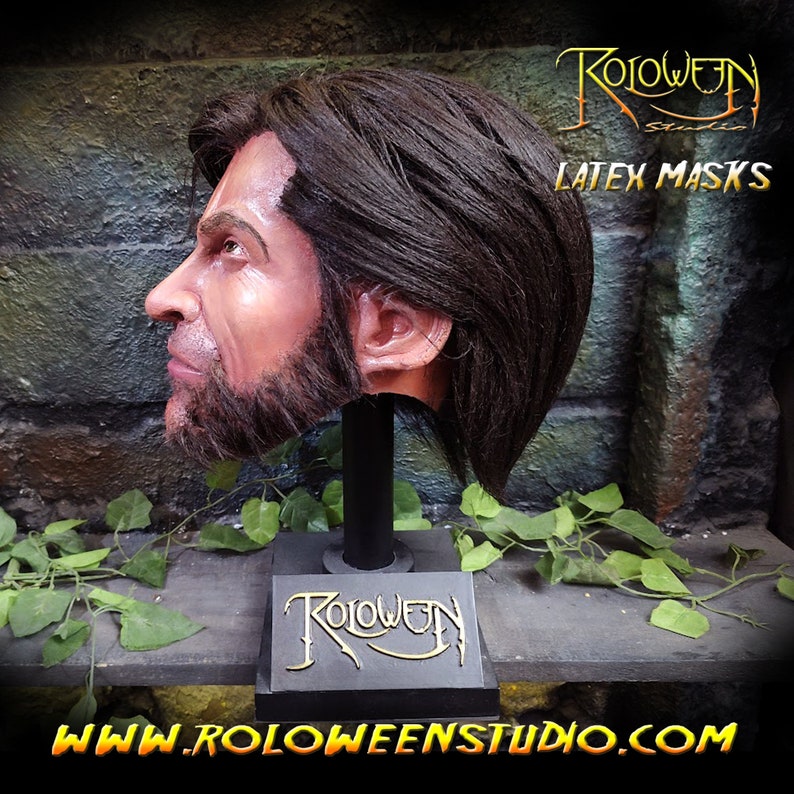 May include: A side profile of a latex mask on a stand. The mask is painted to look like a man with a beard and brown hair. The stand is black with the words "Roloween Studio" on it. The background is a brick wall with green vines.