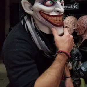 May include: A person wearing a black shirt and blue jeans is holding a green and white Joker mask. The mask has a wide, toothy grin and green hair. The person is also wearing a gray and white striped scarf.