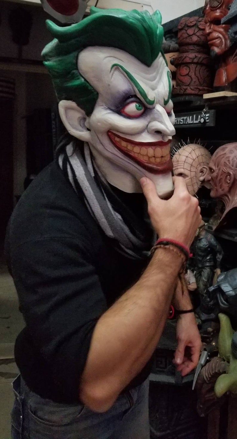 May include: A person wearing a black shirt and blue jeans is holding a green and white Joker mask. The mask has a wide, toothy grin and green hair. The person is also wearing a gray and white striped scarf.