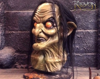 May include: A green latex horror mask with long black hair, a large nose, and glowing orange eyes. The mask is on a wooden stand and has the text "HORROR LATEX MASKS" and "ROLOWEEN" on the background.