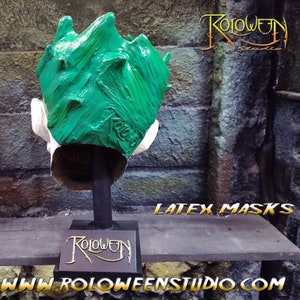May include: Green latex Halloween mask with a textured surface. The mask is on a black stand with a gold logo that says "Roloween Studio". The text "Latex Masks" is visible in the image. The text "www.roloweenstudio.com" is also visible in the image.