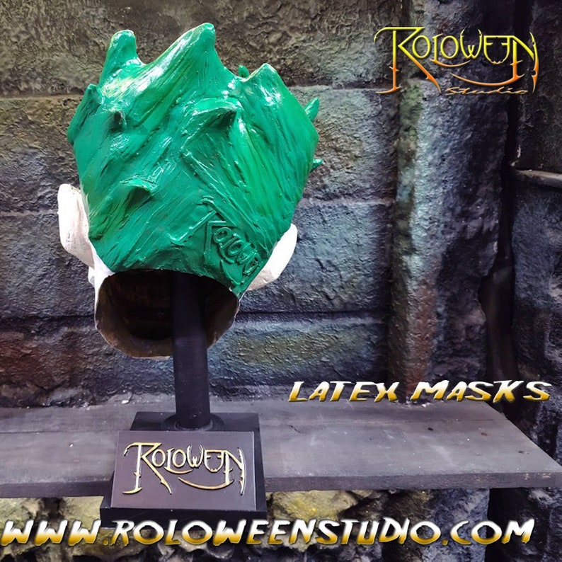 May include: Green latex Halloween mask with a textured surface. The mask is on a black stand with a gold logo that says "Roloween Studio". The text "Latex Masks" is visible in the image. The text "www.roloweenstudio.com" is also visible in the image.