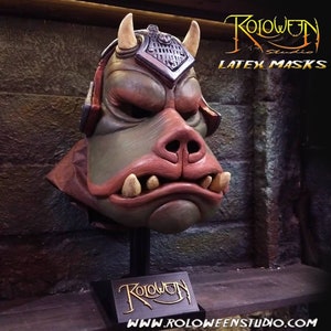 May include: A green and brown latex mask of a creature with large ears, a pointed snout, and sharp teeth. The mask is on a black stand with the text "Roloween Studio" on it. The text "LATEX MASKS" is visible in the upper right corner of the image.