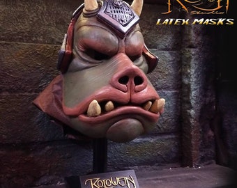 May include: A green and brown latex mask of a creature with large ears, a pointed snout, and sharp teeth. The mask is on a black stand with the text "Roloween Studio" on it. The text "LATEX MASKS" is visible in the upper right corner of the image.
