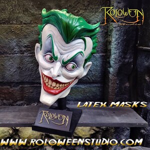 May include: A green and white latex Joker mask with a wide, toothy grin. The mask is on a black stand with the word "Roloween" written on it. The text "Latex Masks" is visible in the background.