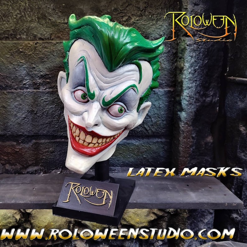 May include: A green and white latex Joker mask with a wide, toothy grin. The mask is on a black stand with the word "Roloween" written on it. The text "Latex Masks" is visible in the background.