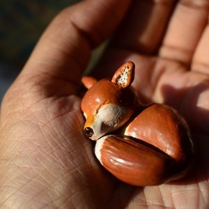 May include: A small, handcrafted figurine of a sleeping fox. The fox is primarily orange with white accents on its belly and ears. The figurine is held in a hand, showcasing its size and detail.