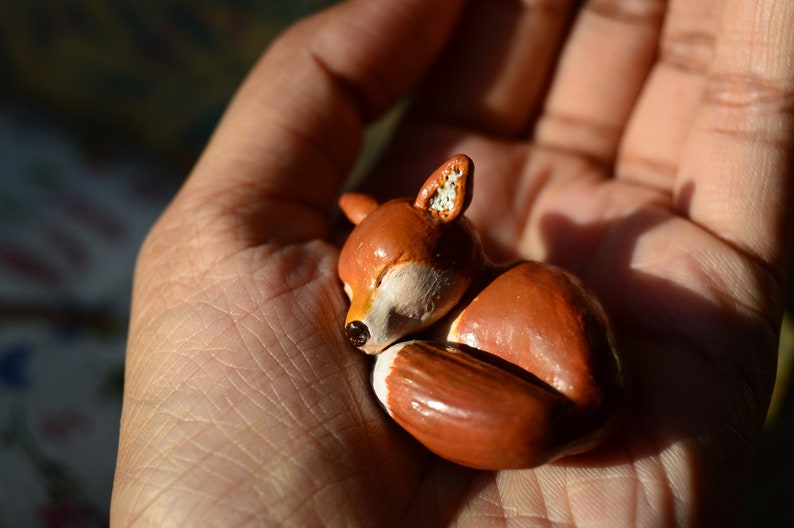May include: A small, handcrafted figurine of a sleeping fox. The fox is primarily orange with white accents on its belly and ears. The figurine is held in a hand, showcasing its size and detail.