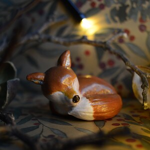 May include: A ceramic figurine of a resting fox, painted in shades of brown and white. The fox is lying down, with its ears perked up. The background features a floral pattern and fairy lights, creating a cozy, woodland theme.