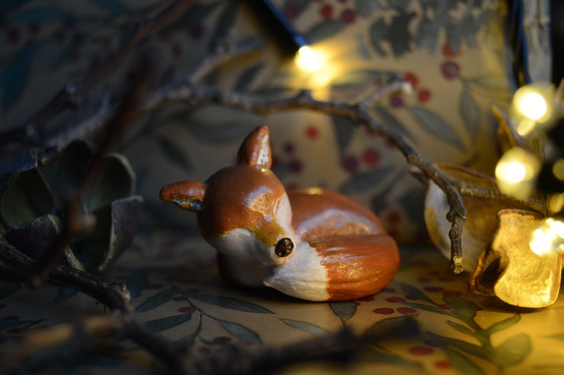 May include: A ceramic figurine of a resting fox, painted in shades of brown and white. The fox is lying down, with its ears perked up. The background features a floral pattern and fairy lights, creating a cozy, woodland theme.