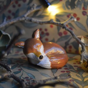 May include: A small, hand-painted ceramic fox figurine in a resting pose. The fox has a brown body, white chest, and a black nose. The figurine is set against a backdrop of a floral pattern and twigs, creating a rustic, woodland theme.