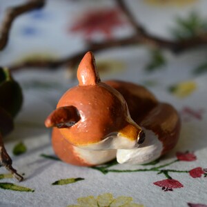 May include: A handcrafted, ceramic fox figurine in a curled-up position. The fox is painted in shades of brown, with white accents on its chest and face. The figurine is set against a floral background with a branch in the background.