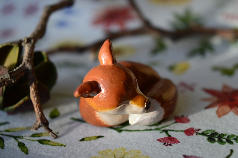May include: A handcrafted, ceramic fox figurine in a curled-up position. The fox is painted in shades of brown, with white accents on its chest and face. The figurine is set against a floral background with a branch in the background.