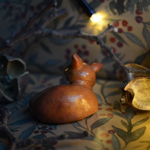 May include: A small, brown ceramic deer figurine with a glossy finish, viewed from the back. The deer is resting on a patterned fabric with botanical motifs. Twigs and dried leaves surround the figurine, with a string of fairy lights in the background.