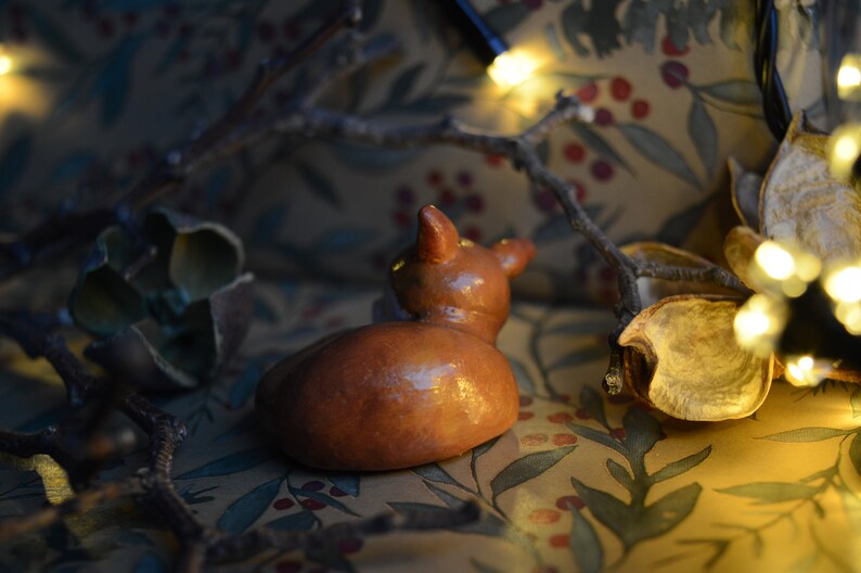 May include: A small, brown ceramic deer figurine with a glossy finish, viewed from the back. The deer is resting on a patterned fabric with botanical motifs. Twigs and dried leaves surround the figurine, with a string of fairy lights in the background.
