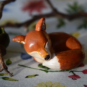 May include: A handcrafted ceramic fox figurine, curled up in a sleeping position. The fox has a reddish-brown coat with a white belly and a black nose. The figurine is set against a floral patterned background with a twig in the upper left corner.