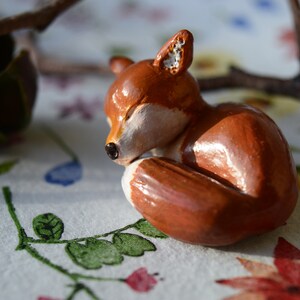 May include: A handcrafted, sleeping fox figurine. The fox is curled up, with its eyes closed, and is painted in shades of brown, white, and black. The figurine is resting on a floral patterned surface, with a branch in the background.