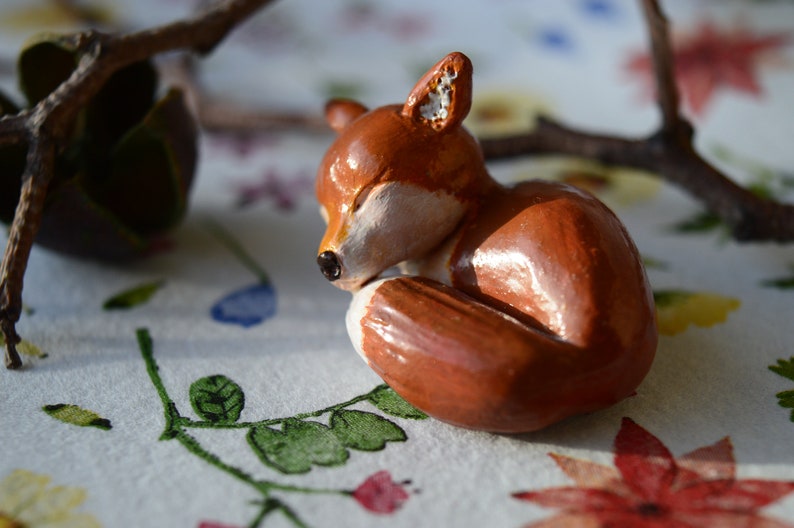 May include: A handcrafted, sleeping fox figurine. The fox is curled up, with its eyes closed, and is painted in shades of brown, white, and black. The figurine is resting on a floral patterned surface, with a branch in the background.