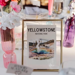 May include: A framed print with the text "YELLOWSTONE NATIONAL PARK" and a watercolor image of a geyser and surrounding landscape. The frame is gold, and the print is displayed on a white table, with floral arrangements in the background.