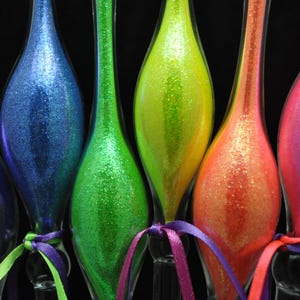 May include: Six colorful, glitter-covered glass ornaments in a row. The ornaments are in shades of purple, blue, green, yellow, orange, and pink. Each ornament is tied with a matching ribbon. The background is black.