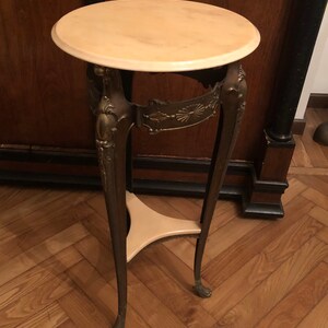 May include: A round, cream-coloured marble top table with a gold-toned metal base. The base has intricate details and three legs.