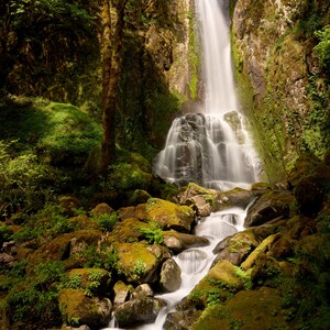 May include: A tall waterfall cascades down a rocky cliff face into a stream flowing over moss-covered boulders. The scene is surrounded by dense green forest foliage and trees. The text Photography by Ffej is visible at the bottom.